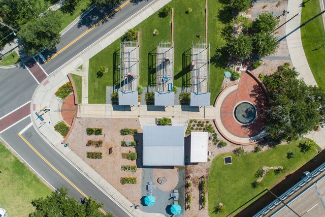 An aerial top-down view of Smokey Hollow Commemorative Park, featuring three open-frame house outline sculptures on green lawn, a brick circular plaza with a fountain, a central pavilion building, garden plots, and paved pedestrian walkways situated next to city streets.
