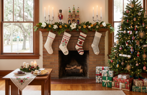 Share your Christmas traditions with us A neatly organized, bright living room decorated for Christmas, featuring a blend of global traditions. A brick fireplace is central, adorned with magnolia leaves, dried citrus slices, white candles, a wooden nutcracker, and a traditional German Christmas pyramid (Pyramide). Four distinct embroidered knit stockings hang from the mantel. To the right stands an evergreen tree decorated with straw stars, glass ornaments, Ukrainian-style painted eggs, and British Christmas crackers, surrounded by wrapped gifts. On a wooden coffee table in the foreground sits an Advent wreath with four lit white candles and plates of traditional holiday cookies. Large windows show a sunny, tree-lined view outside.