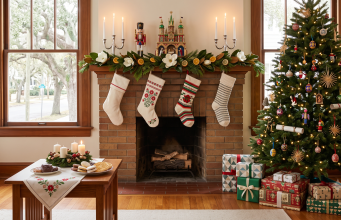 Share your Christmas traditions with us A neatly organized, bright living room decorated for Christmas, featuring a blend of global traditions. A brick fireplace is central, adorned with magnolia leaves, dried citrus slices, white candles, a wooden nutcracker, and a traditional German Christmas pyramid (Pyramide). Four distinct embroidered knit stockings hang from the mantel. To the right stands an evergreen tree decorated with straw stars, glass ornaments, Ukrainian-style painted eggs, and British Christmas crackers, surrounded by wrapped gifts. On a wooden coffee table in the foreground sits an Advent wreath with four lit white candles and plates of traditional holiday cookies. Large windows show a sunny, tree-lined view outside.