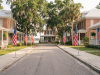 How Flag Day got its start A wide, eye-level photograph looking down a quiet, sunlit residential street lined with historic brick and wooden-siding homes. Large American flags on temporary metal stands line both sides of the concrete sidewalk, leading toward the end of the road. Additional American flags and patriotic bunting hang from the front porches of the houses. Spanish moss drapes from mature oak and palm trees framing the quiet asphalt street under a bright sky.