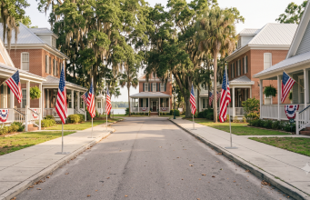 How Flag Day got its start A wide, eye-level photograph looking down a quiet, sunlit residential street lined with historic brick and wooden-siding homes. Large American flags on temporary metal stands line both sides of the concrete sidewalk, leading toward the end of the road. Additional American flags and patriotic bunting hang from the front porches of the houses. Spanish moss drapes from mature oak and palm trees framing the quiet asphalt street under a bright sky.