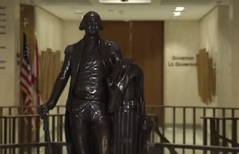 Leon County invites residents to rediscover local history during America 250 celebration A dark bronze full-body statue of George Washington in military uniform holding a walking cane, standing in a brightly lit government building corridor. In the background, an American flag and a state flag stand next to a hallway wall bearing the text 'Governor' and 'Lt. Governor'.