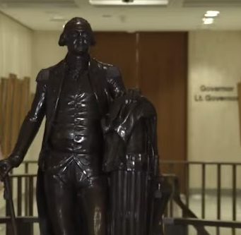 Leon County invites residents to rediscover local history during America 250 celebration A dark bronze full-body statue of George Washington in military uniform holding a walking cane, standing in a brightly lit government building corridor. In the background, an American flag and a state flag stand next to a hallway wall bearing the text 'Governor' and 'Lt. Governor'.
