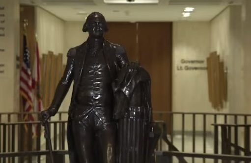 Leon County invites residents to rediscover local history during America 250 celebration A dark bronze full-body statue of George Washington in military uniform holding a walking cane, standing in a brightly lit government building corridor. In the background, an American flag and a state flag stand next to a hallway wall bearing the text 'Governor' and 'Lt. Governor'.