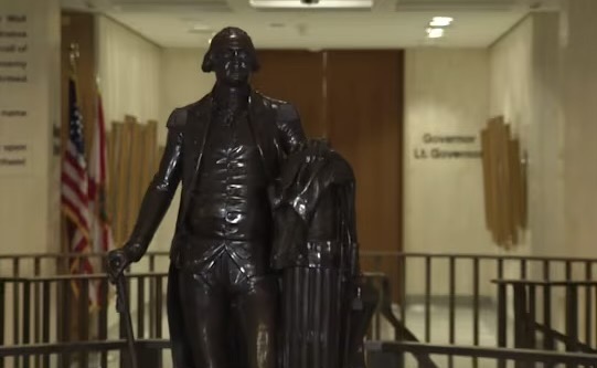 history A dark bronze full-body statue of George Washington in military uniform holding a walking cane, standing in a brightly lit government building corridor. In the background, an American flag and a state flag stand next to a hallway wall bearing the text 'Governor' and 'Lt. Governor'.